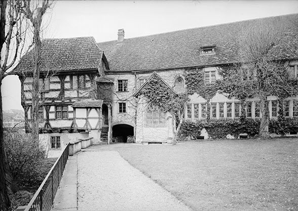 View over a lawn of a building with two floors. There is one tree in front of the building. The walls are ivy-clad. The ground-floor windows form a continuous row. On the left is a pavilion-like half-timbered structure. The photo is in black and white.